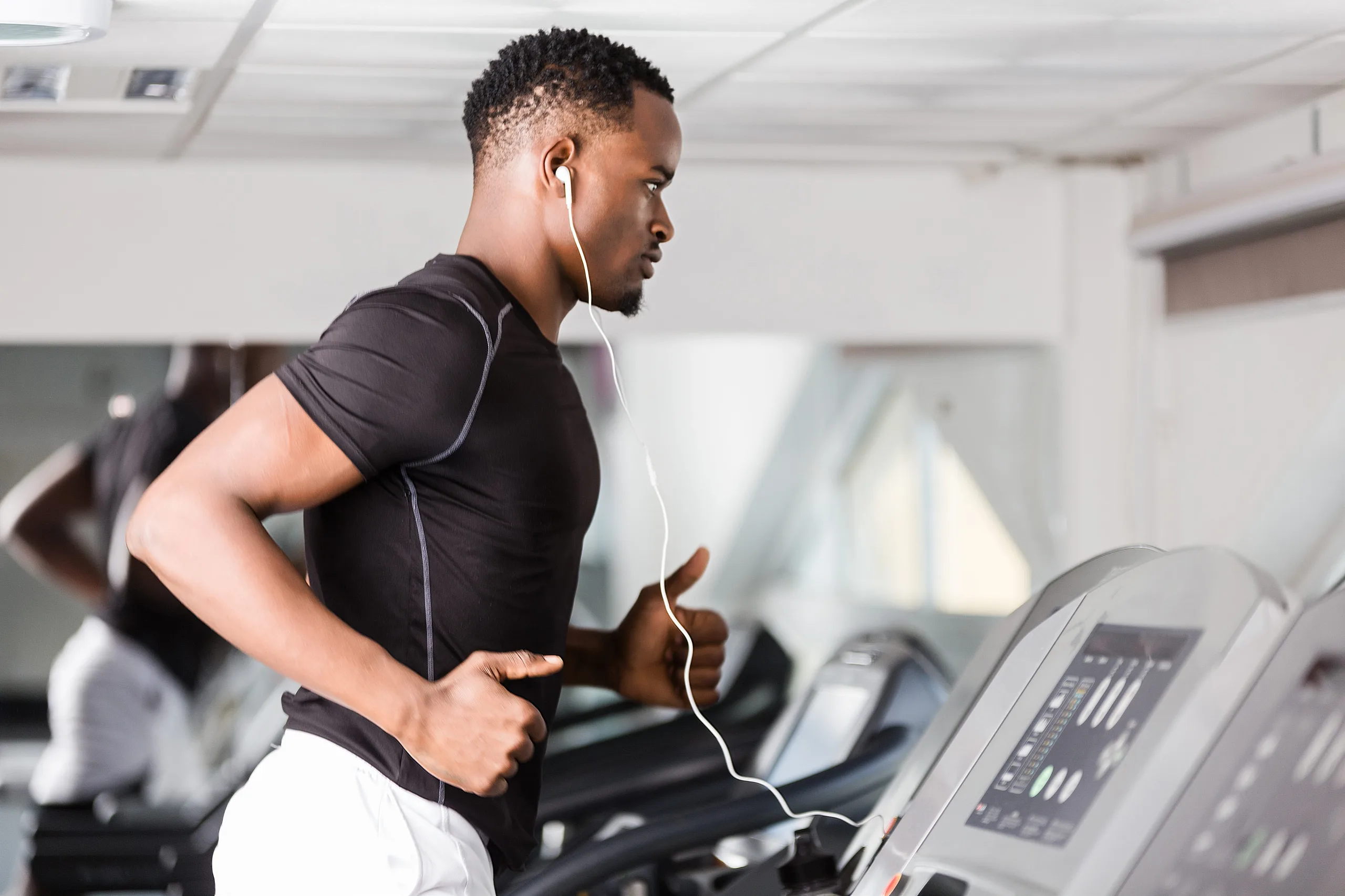 Man running on a treadmill with his earphones wired into the machine. he looks focused and is wearing a tight black shirt and light grey shorts on.