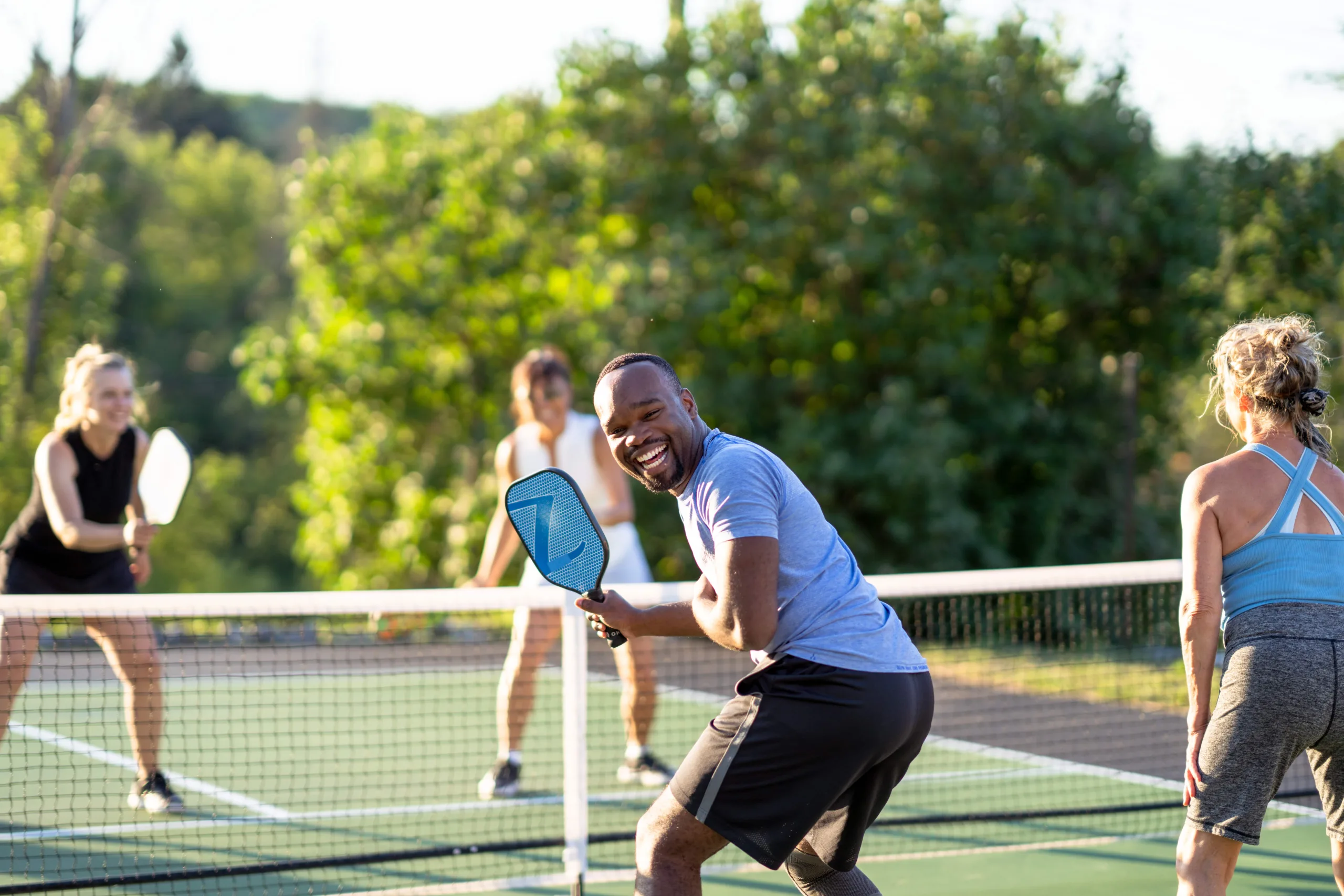 Man in Black shorts and athletic top with a pickleball racket in his hand looking back smiling at the camera while his partner looks forward as another player is getting ready to serve the ball. The background is blurred and hosts vibrant foliage and trees on a hillside.