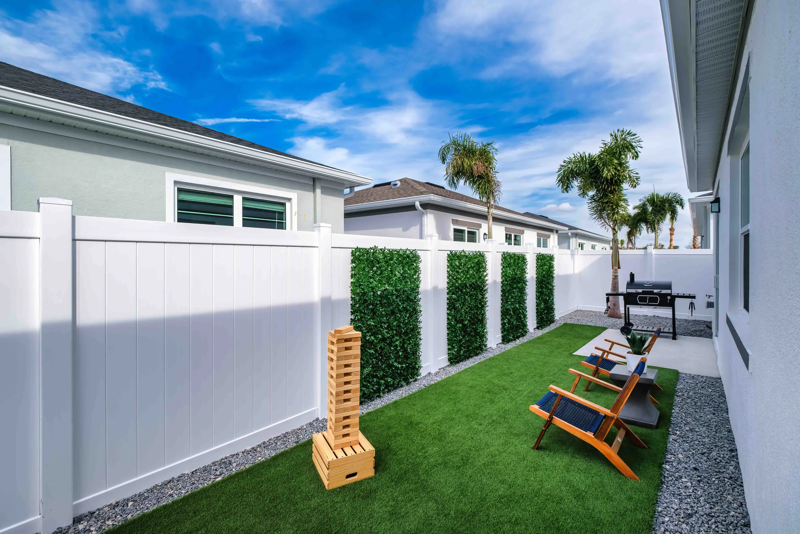 A beautiful Florida backyard scaped with artificial turf, a mature palm tree, seating, a grill, a patio, and a white fence on a beautiful breezy sunny day.