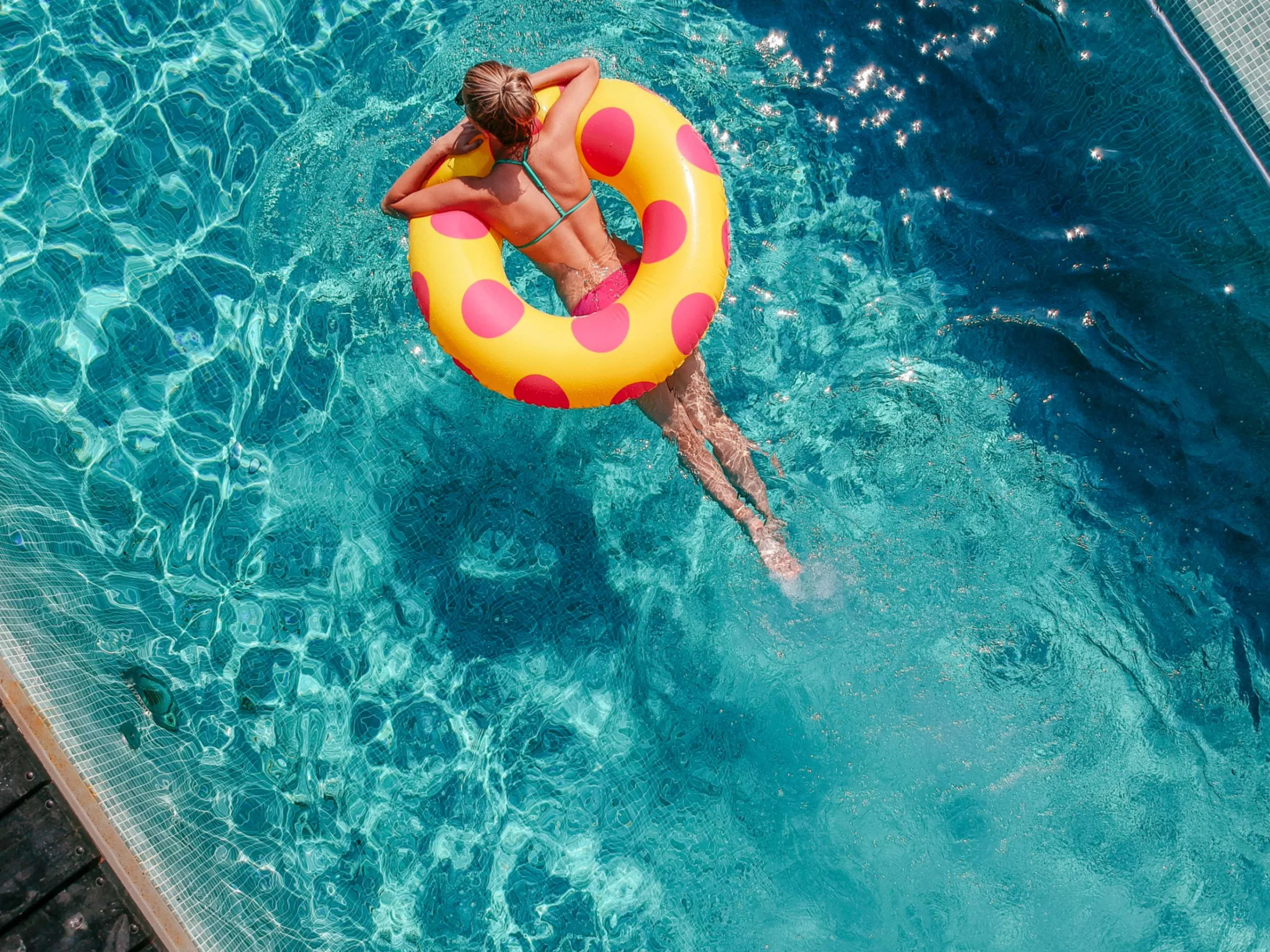 Woman relaxing in a pool with an inflatable tube that's yellow with pink polka dots. She has blonde hair in a bun and sunglasses on.